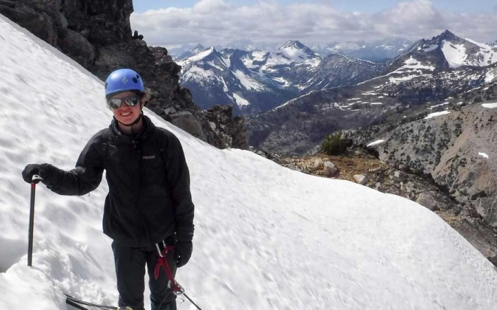 A person wearing safety gear stands on a snowy incline smiling at the camera. Behind them, there are vast, snow-covered mountains.